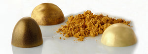 tre praline di cioccolato nei colori oro chiaro, oro scuro e perlato accanto a polvere dorata sfusa. Tutto posizionato su un piano bianco lucido