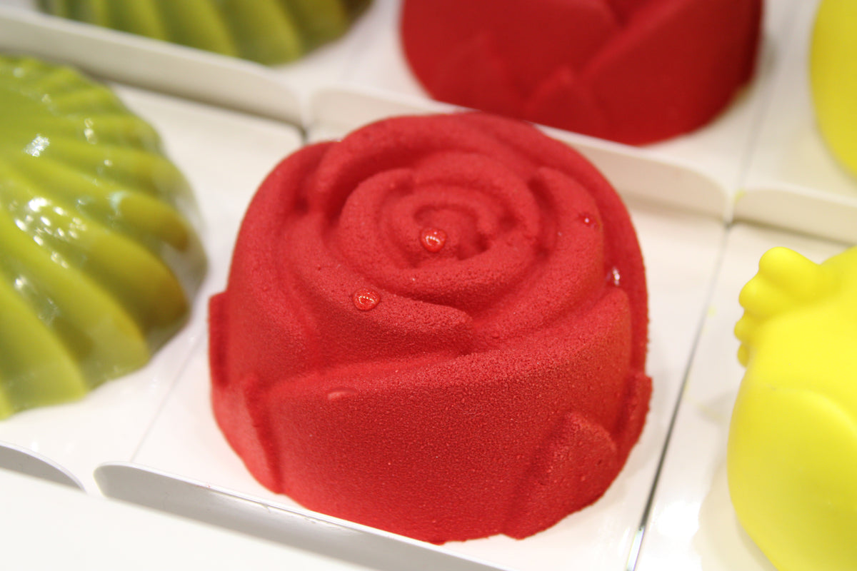 Piccolo tortino a forma di rosa rossa con superficie ad effetto velluto su base in cartoncino bianco. Decorazioni a goccia trasparenti sui petali. Sullo sfondo altre piccole torte di forme e colori diversi.
