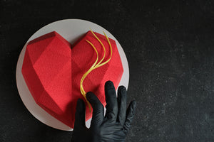 Dolce a forma di cuore rosso effetto vellutato con superficie sfaccettata come un diamante su piatto bianco e sfondo nero.
Una mano con guanto nero posiziona al centro del cuore una decorazione a tre fili di cioccolato di colore giallo 