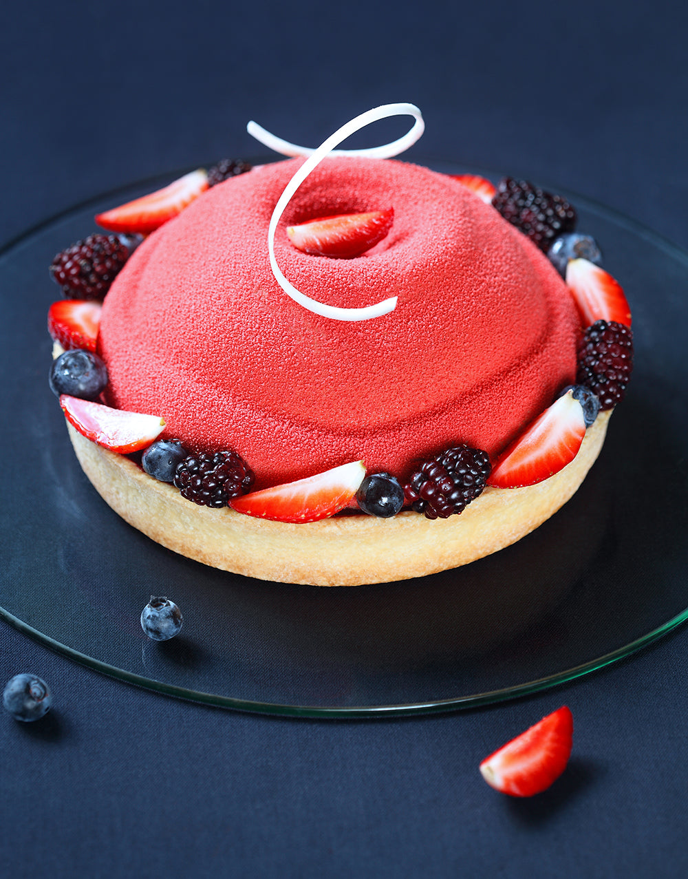 torta tonda su piatto di vetro sfondo blu. Torta con effetto velluto rosso con base di pasta frolla decorata con frutti di bosco vari.