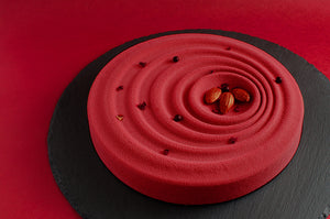 Torta tonda rossa effetto velluto con superficie ad onde concentriche su base di pietra nera e sfondo rosso. Piccole applicazioni decorative e tre mandorle dorate.