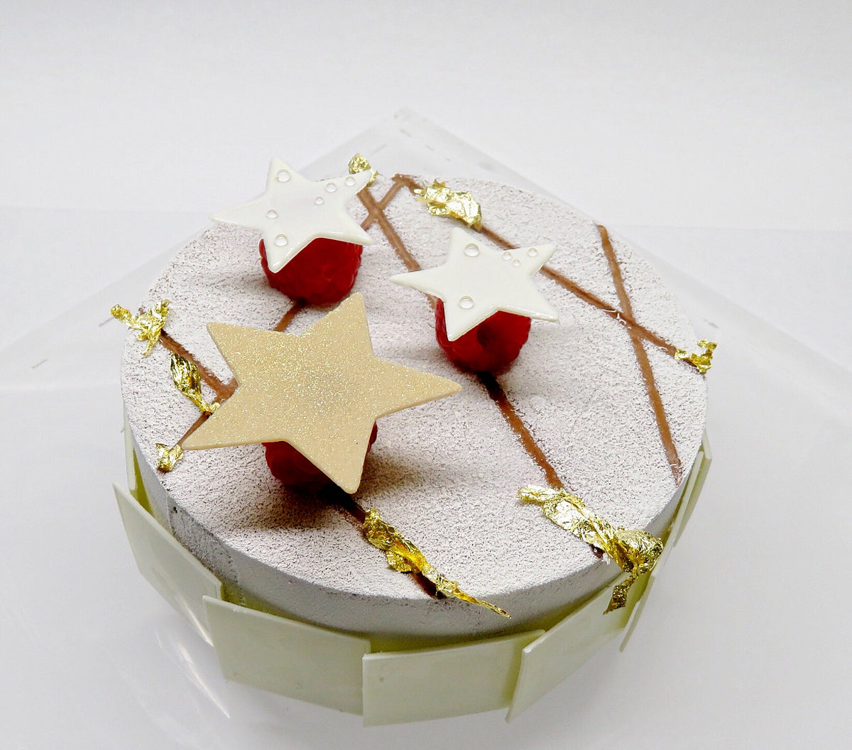 Torta bianca con bordo rivestito di quadretti di cioccolato bianco. Effetto vellutato bianco in superficie, con decorazione di foglietti d'oro, 3 lamponi sormontati da tre stelle di cioccolato bianco di cui una dorata.